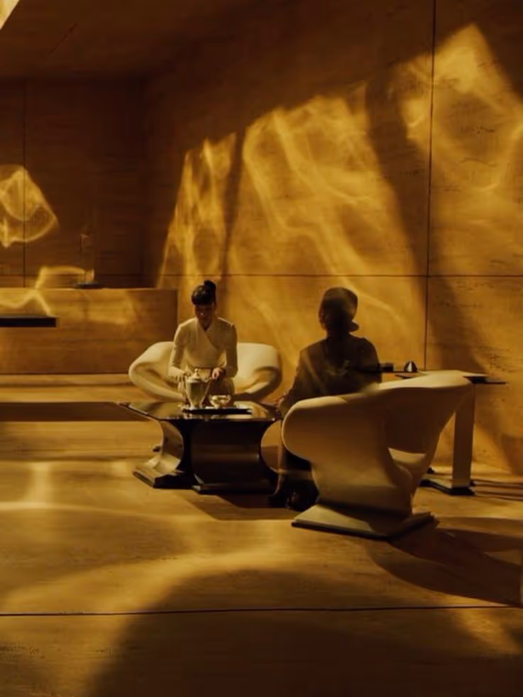 Two people seated in white modern chairs around a black table with golden light patterns on walls and floor.
