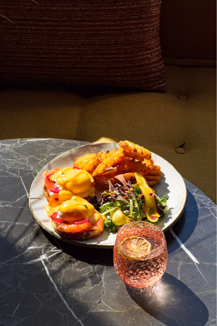 Plate with two eggs benedict topped with hollandaise sauce, waffle fries, and mixed salad, accompanied by a pink textured glass with a lemon slice on a dark marble table.