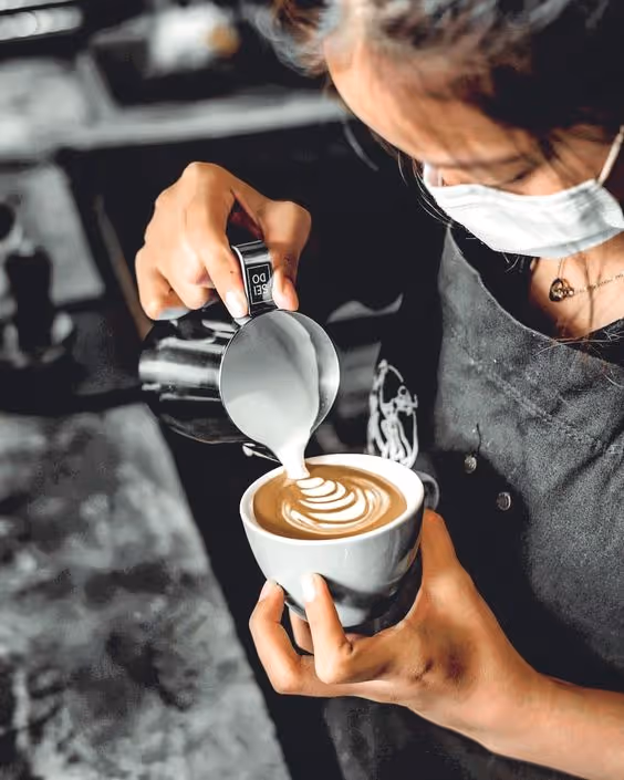 Barista wearing a mask pouring steamed milk to create latte art in a white cup.