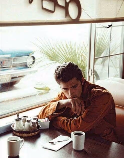 Man in a brown jacket sitting at a cafe table with two coffee mugs, looking thoughtfully out the window.