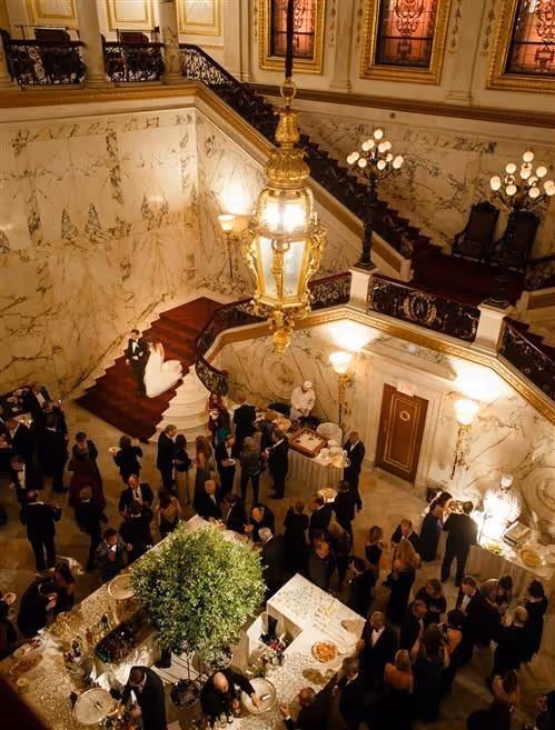 Overhead view of a formal gathering with guests mingling around tables in a grand marble hall with a large chandelier and stained glass windows, and a bride and groom sitting on stairs.