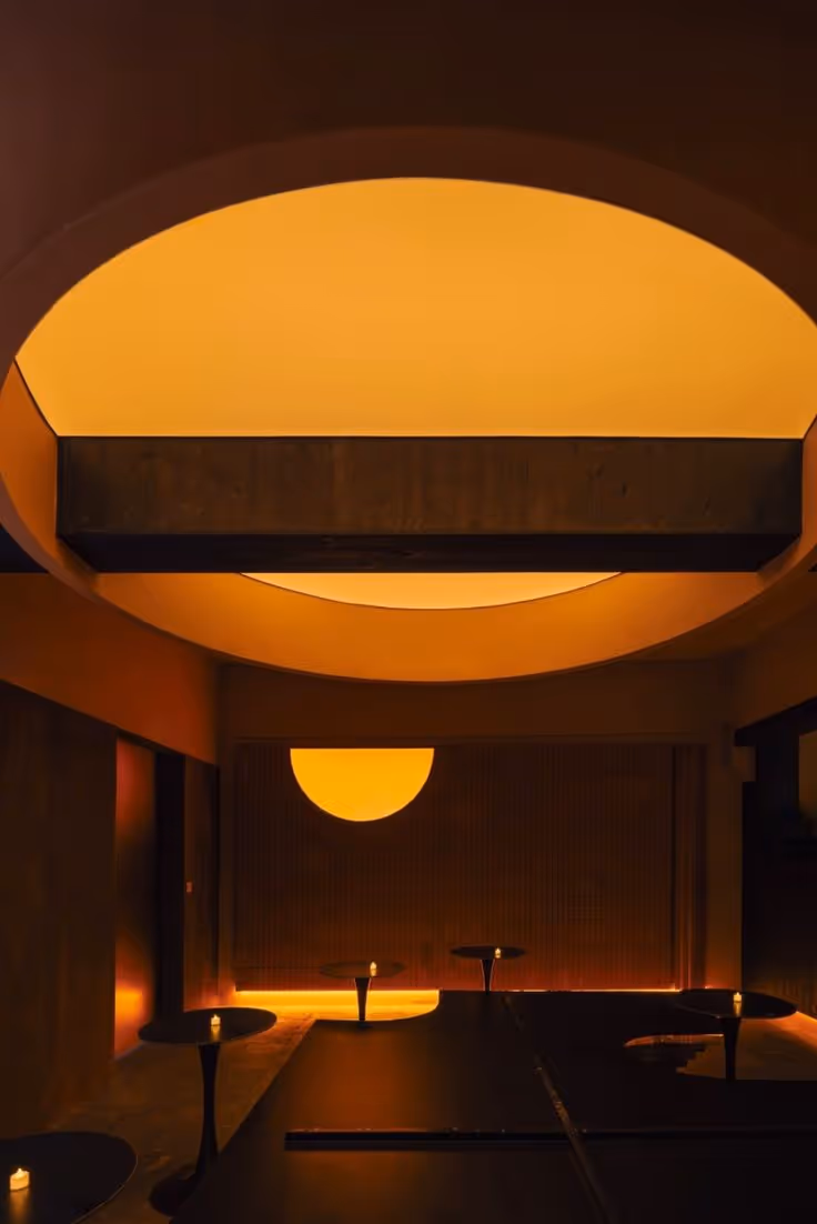 Dimly lit room with a large circular ceiling light and small candles on round tables creating a warm amber glow.