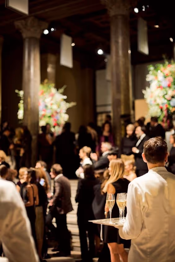 Waiter in white uniform holding a tray with champagne glasses at an elegant indoor event with many well-dressed guests.