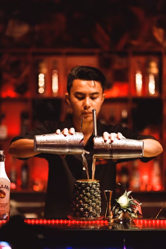 Bartender pouring a cocktail from two shakers into a hollowed-out pineapple at a bar with red lighting.