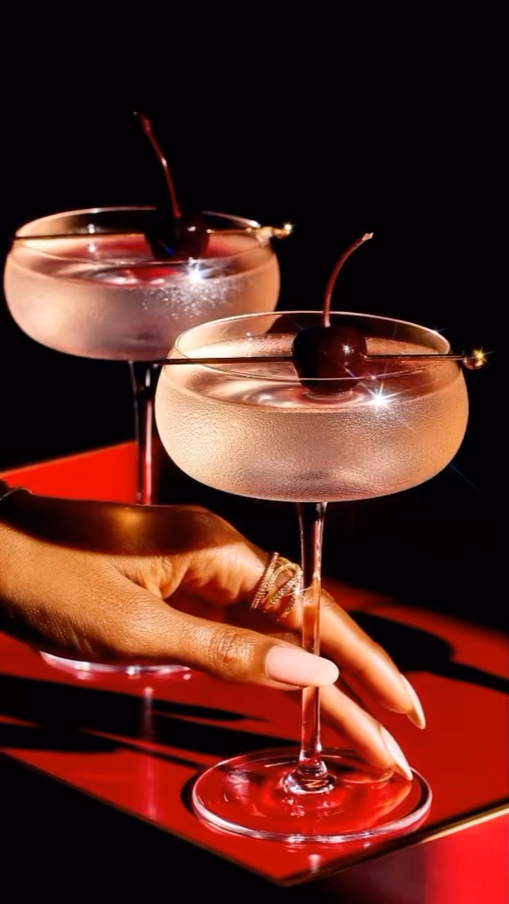 Hand with gold rings holding a cocktail glass garnished with a cherry on a red surface, with another similar glass in the background.