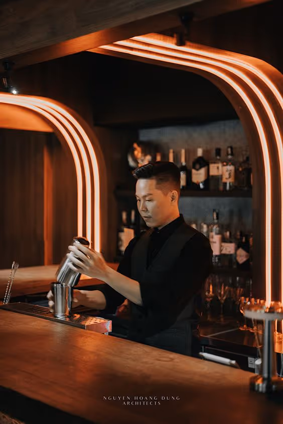 Bartender pouring a drink behind a bar with modern orange neon arch lights and shelves of liquor bottles in the background.