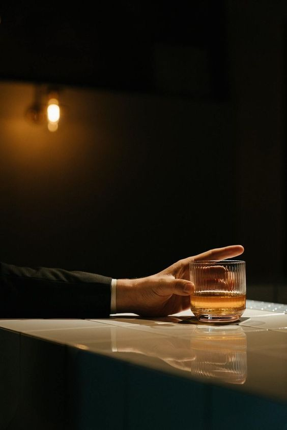 Hand wearing a dark suit resting on a bar counter while holding a glass of whiskey under warm lighting.
