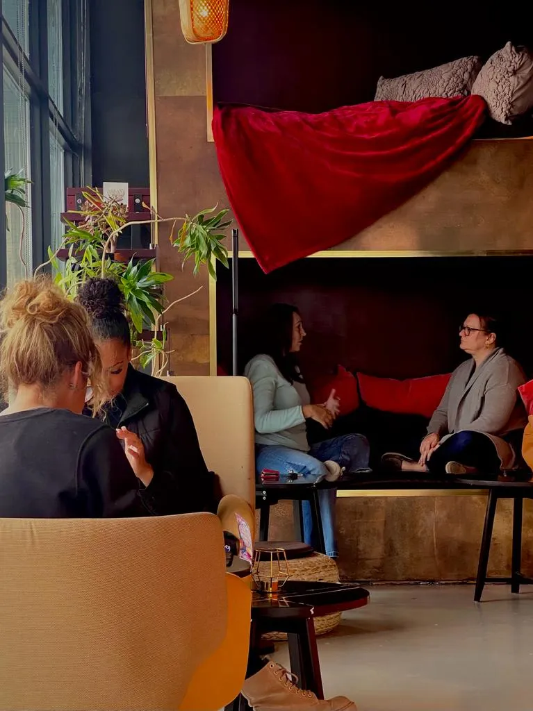 Four people sitting and chatting in a cozy room with plants, a red blanket, and cushions.