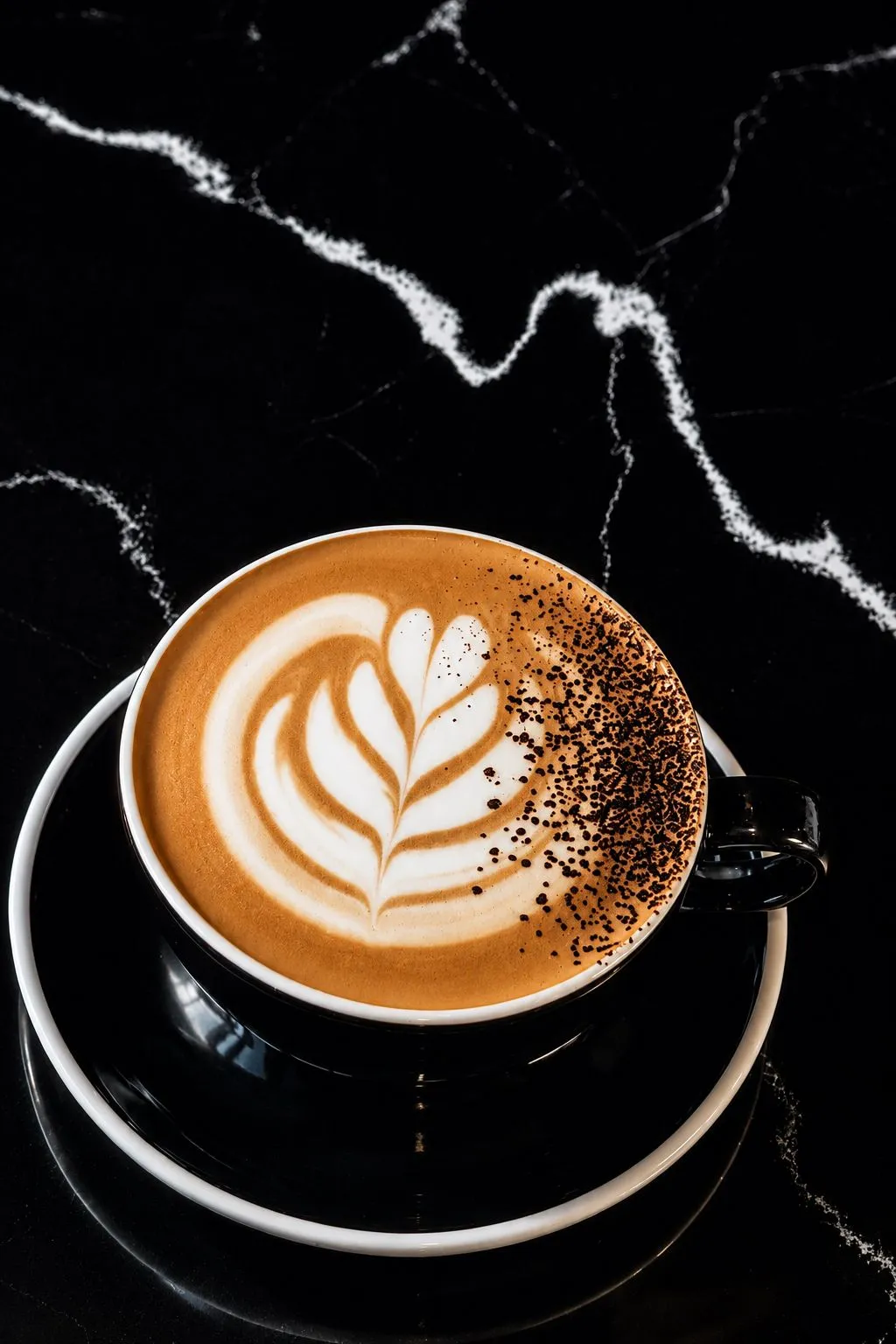 Black coffee cup with saucer containing latte with leaf-shaped foam art and sprinkled chocolate.