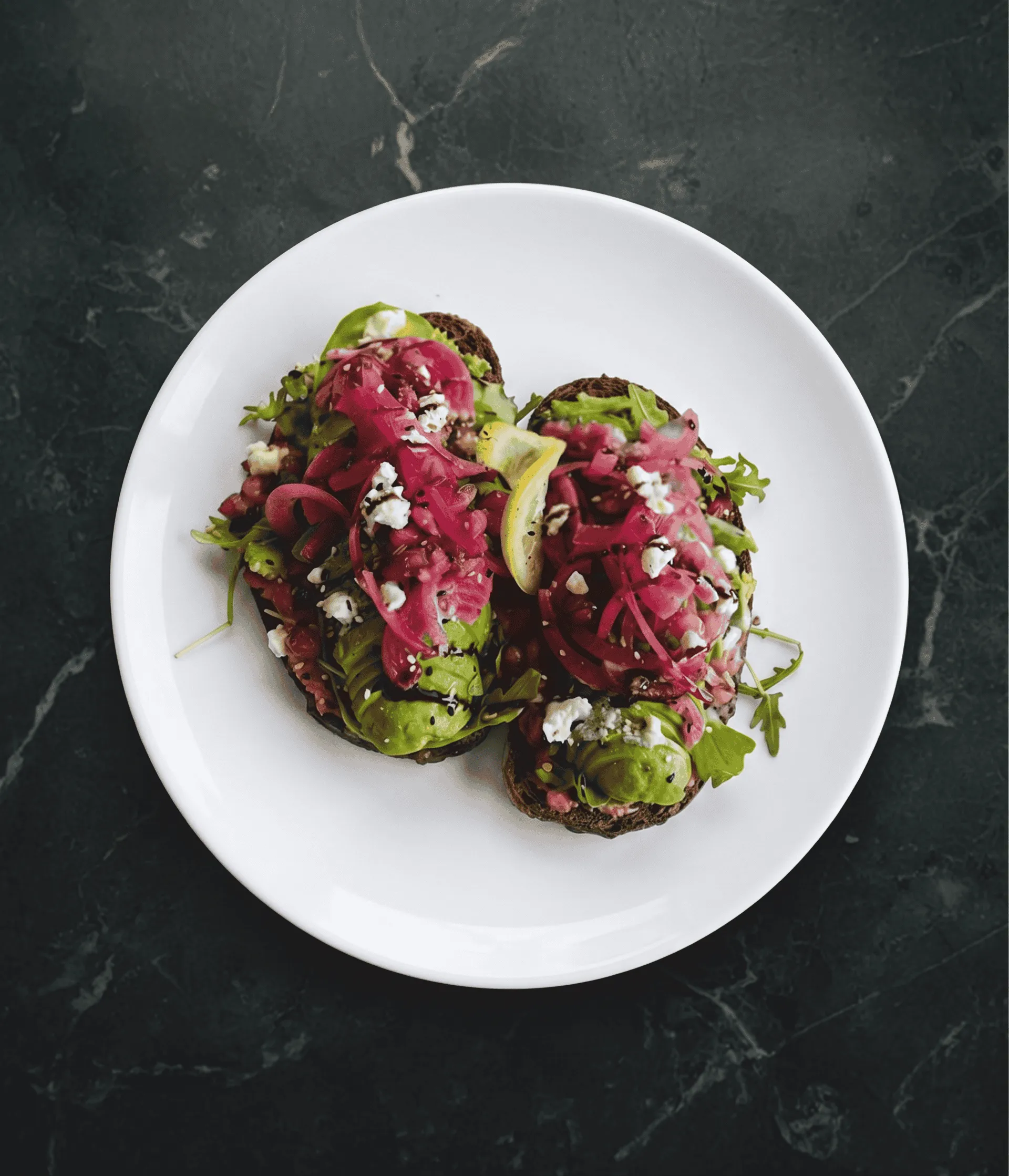 Two avocado toast slices topped with pickled onions, feta, greens, and a lemon wedge on a white plate.