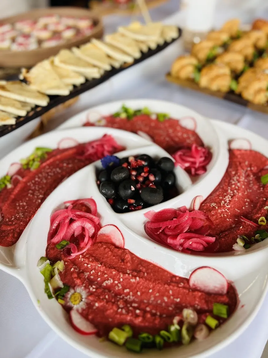 White divided dish with red beet hummus garnished with onions, radishes, green onions, and black olives.