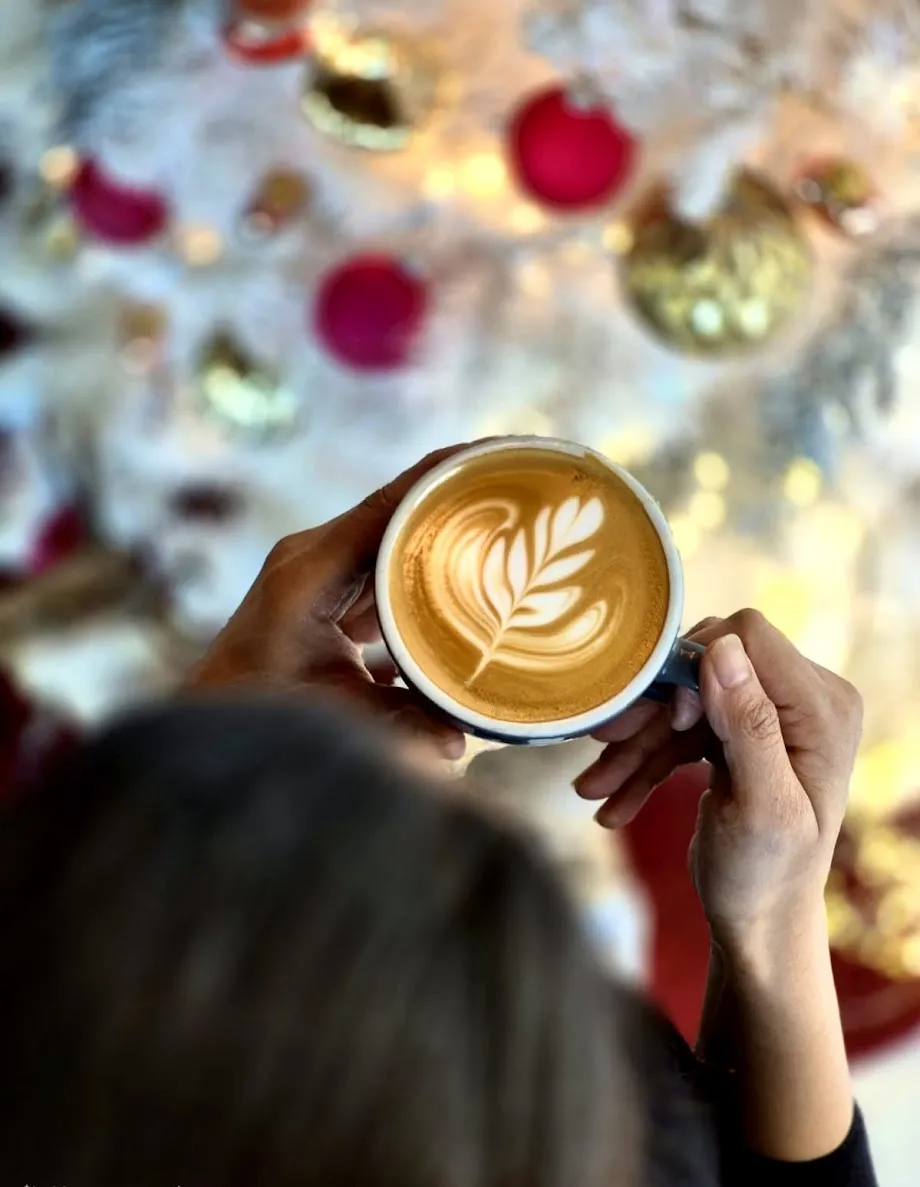 Person holding a cup of latte with leaf latte art, blurred festive decorations in background.