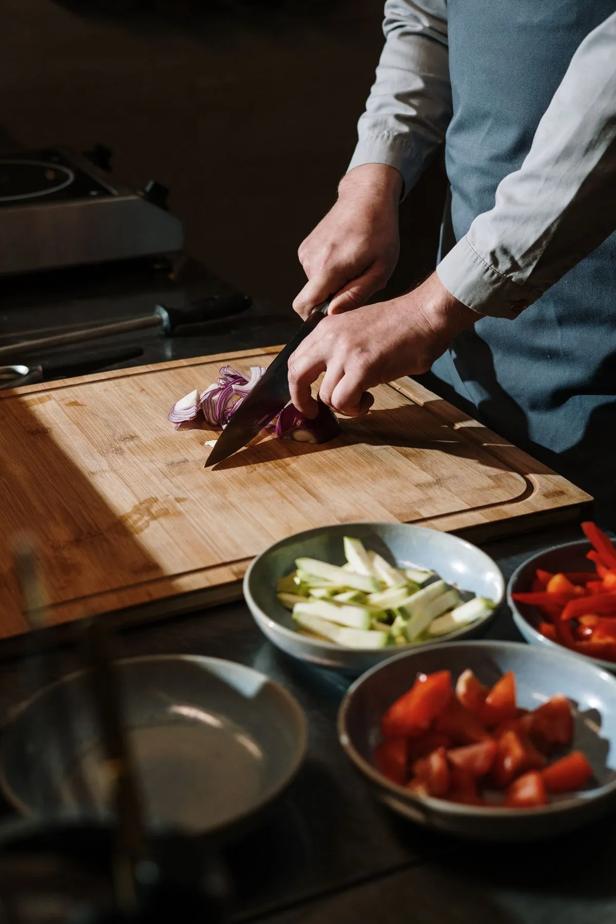 Person slicing a red onion on a wooden cutting board with bowls of chopped vegetables nearby.