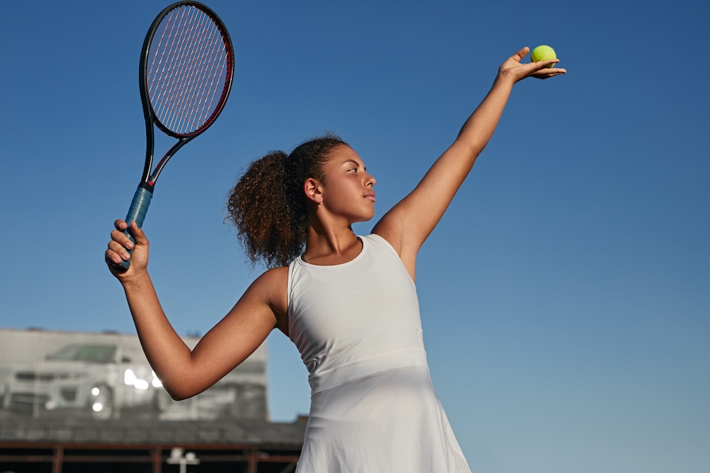 Tennis player in white dress preparing to serve ball against blue sky