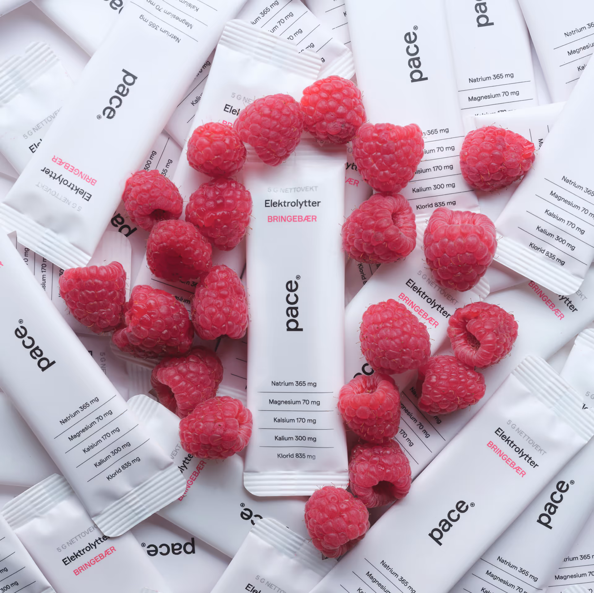 White electrolyte powder packets labeled 'pace' and 'Bringebær' surrounded by fresh raspberries.