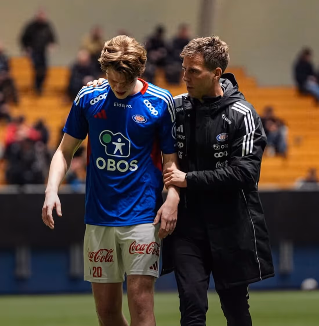 A soccer coach wearing a black jacket supports a player in a blue jersey and white shorts on the field.