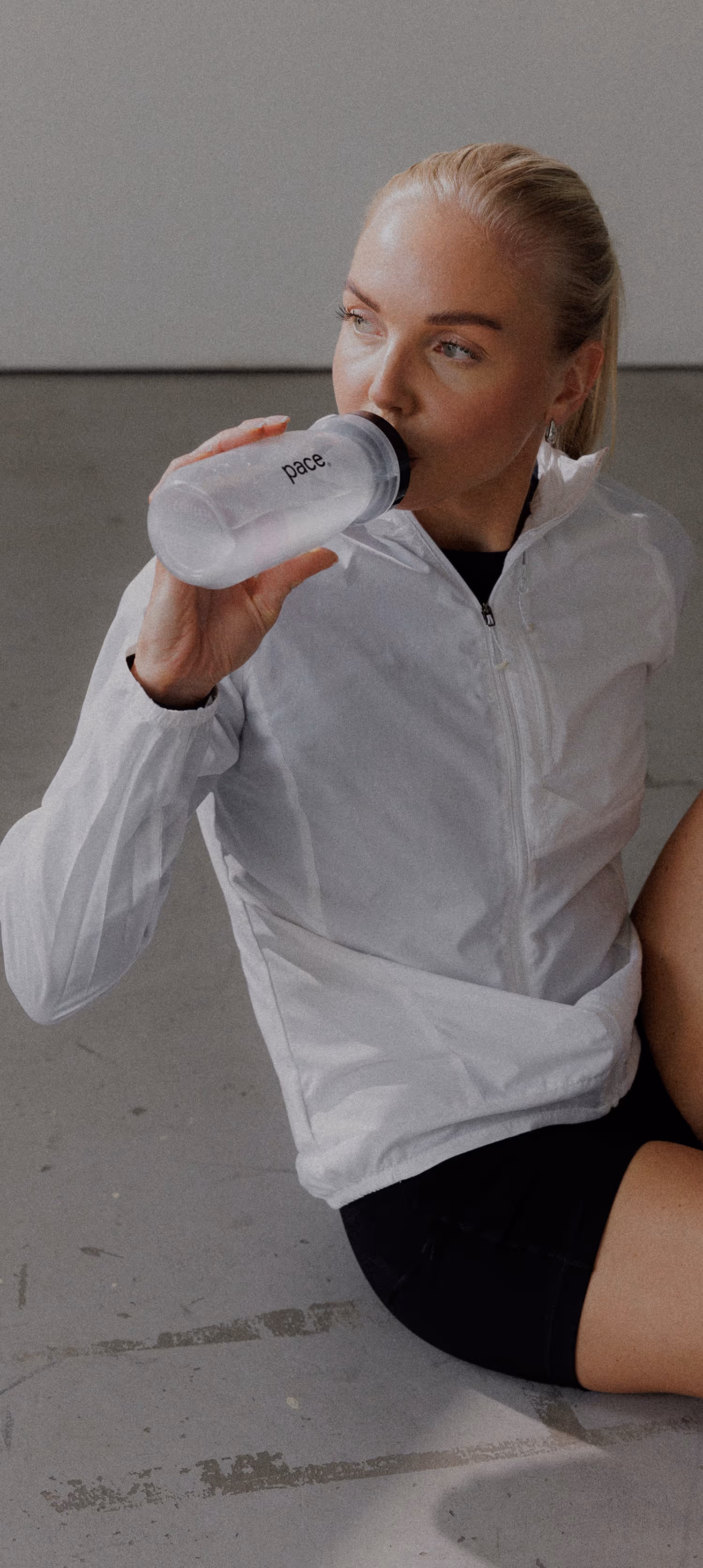 Blonde woman in a white jacket sitting on the floor and drinking water from a bottle labeled 'pace.'