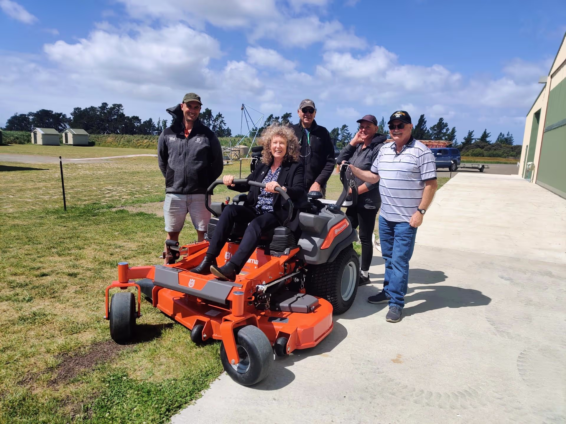 Group of people gathered around a person seated on an orange zero-turn lawnmower.