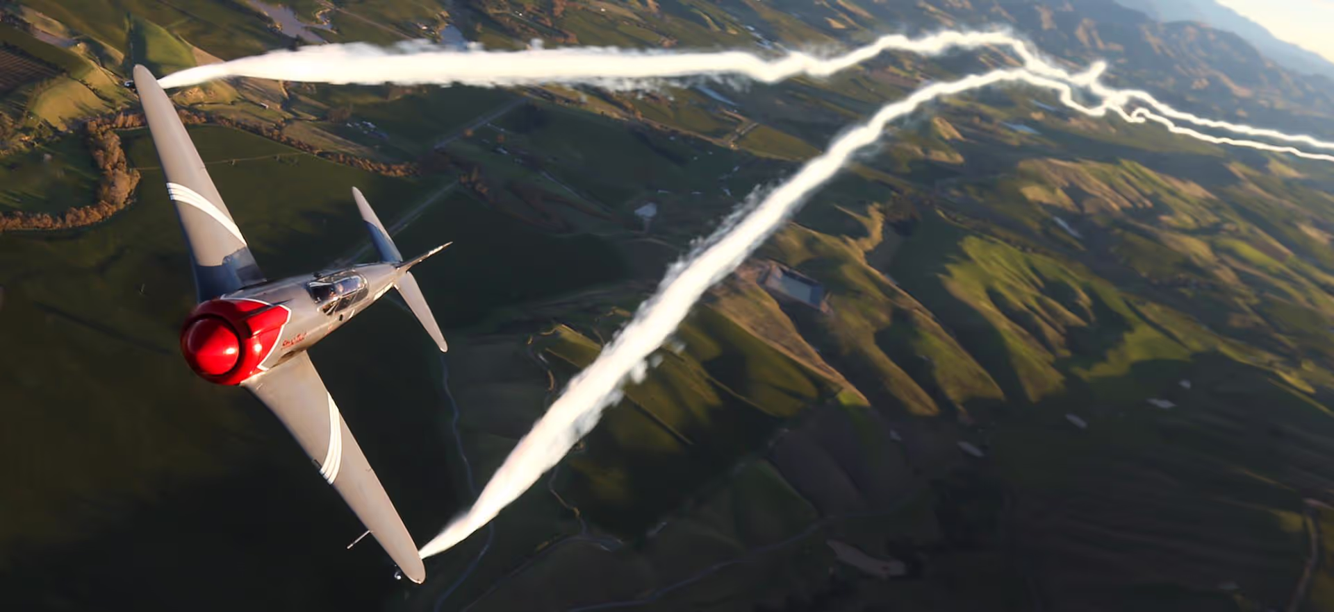 Two gliders flying in close formation above a green landscape, leaving white smoke trails.