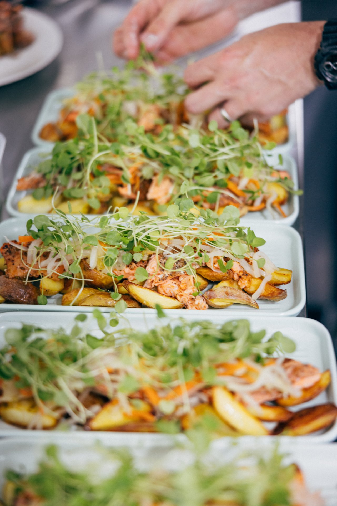 Hands garnishing trays of salmon and potatoes with fresh sprouts.