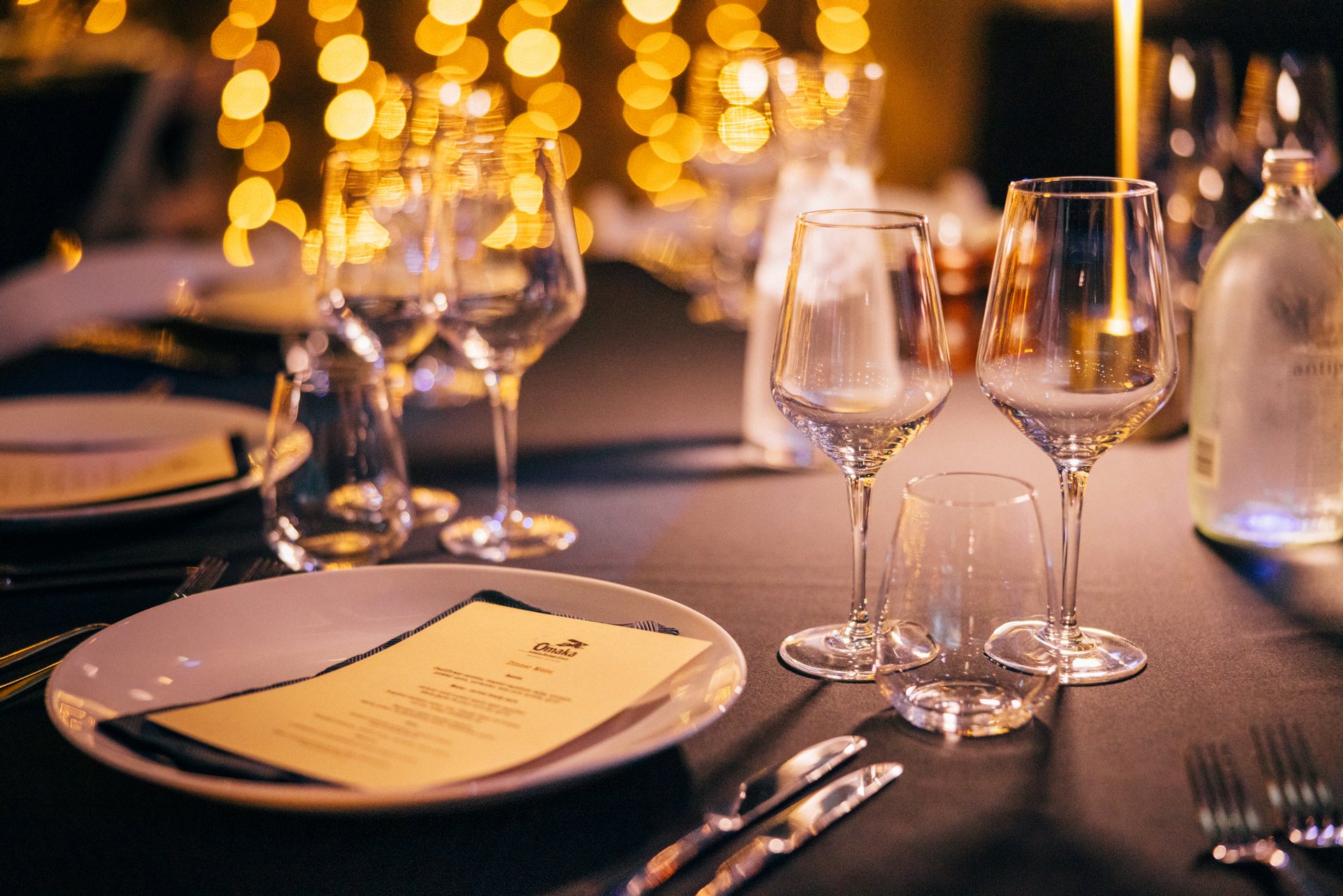 Elegant dinner setting with glasses and menu on a table, warm bokeh lights in background.