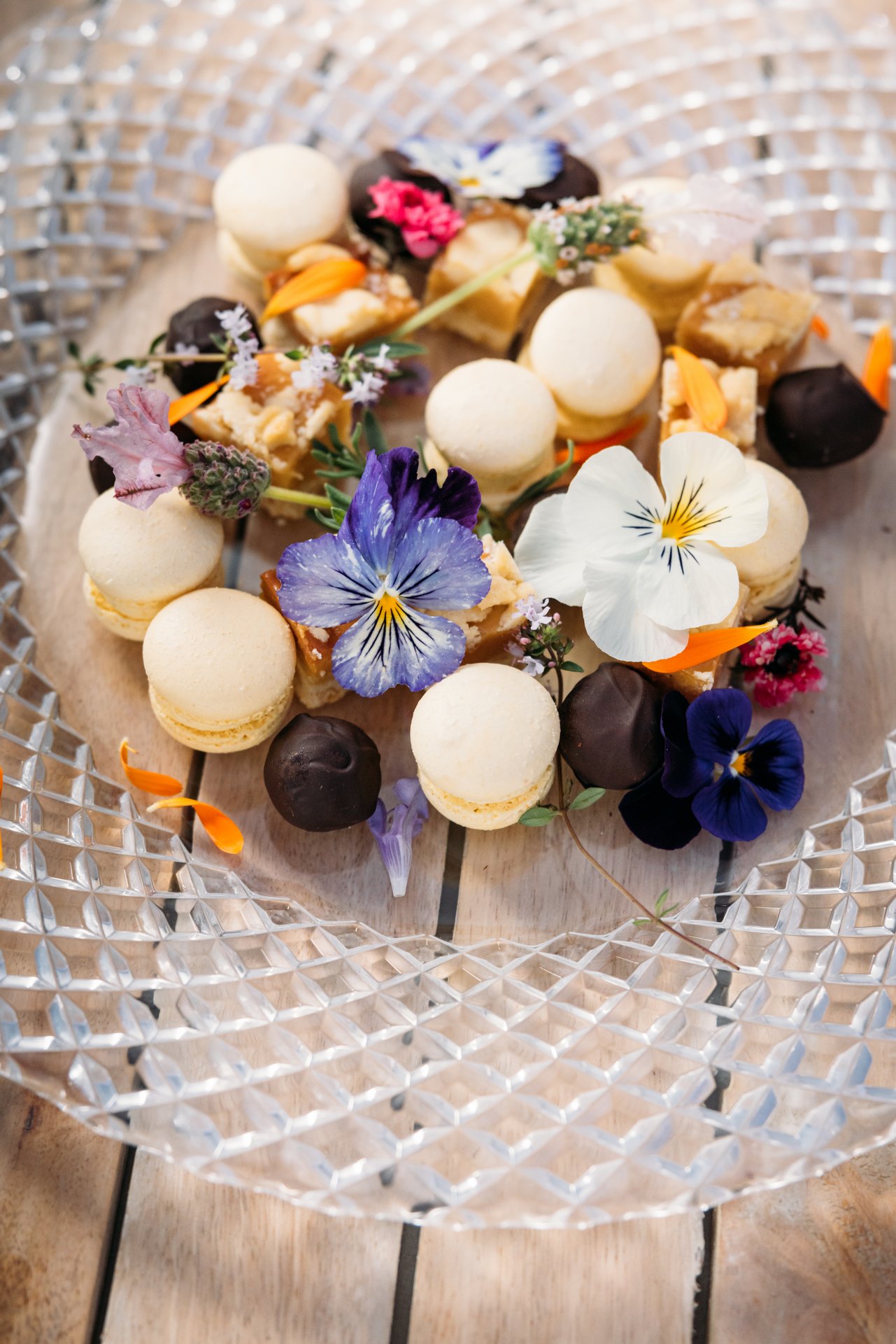 Assorted macarons, chocolates, and edible flowers on a wooden board with a crystal tray.