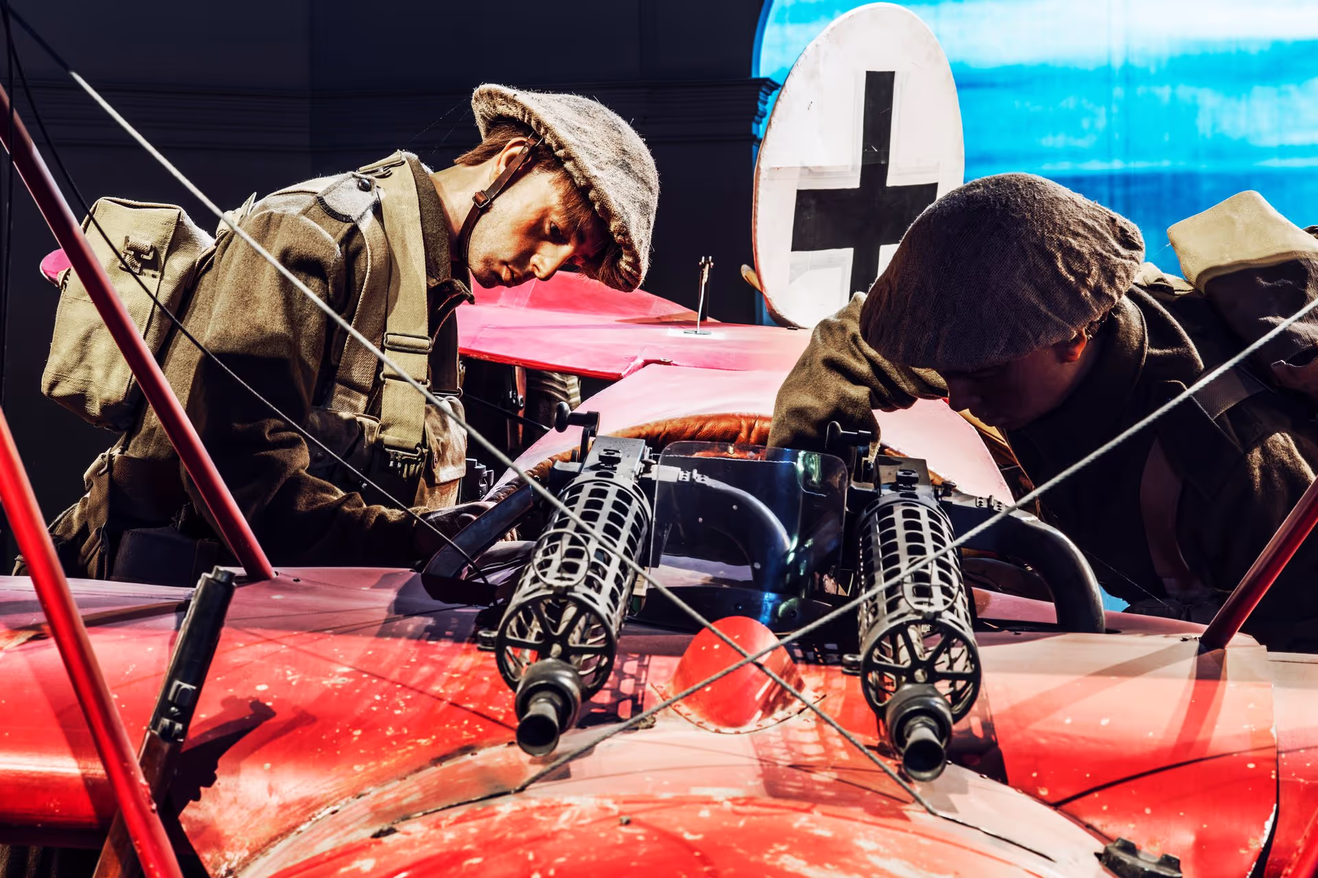 Two figures in vintage pilot gear working on a red biplane with a white cross emblem.