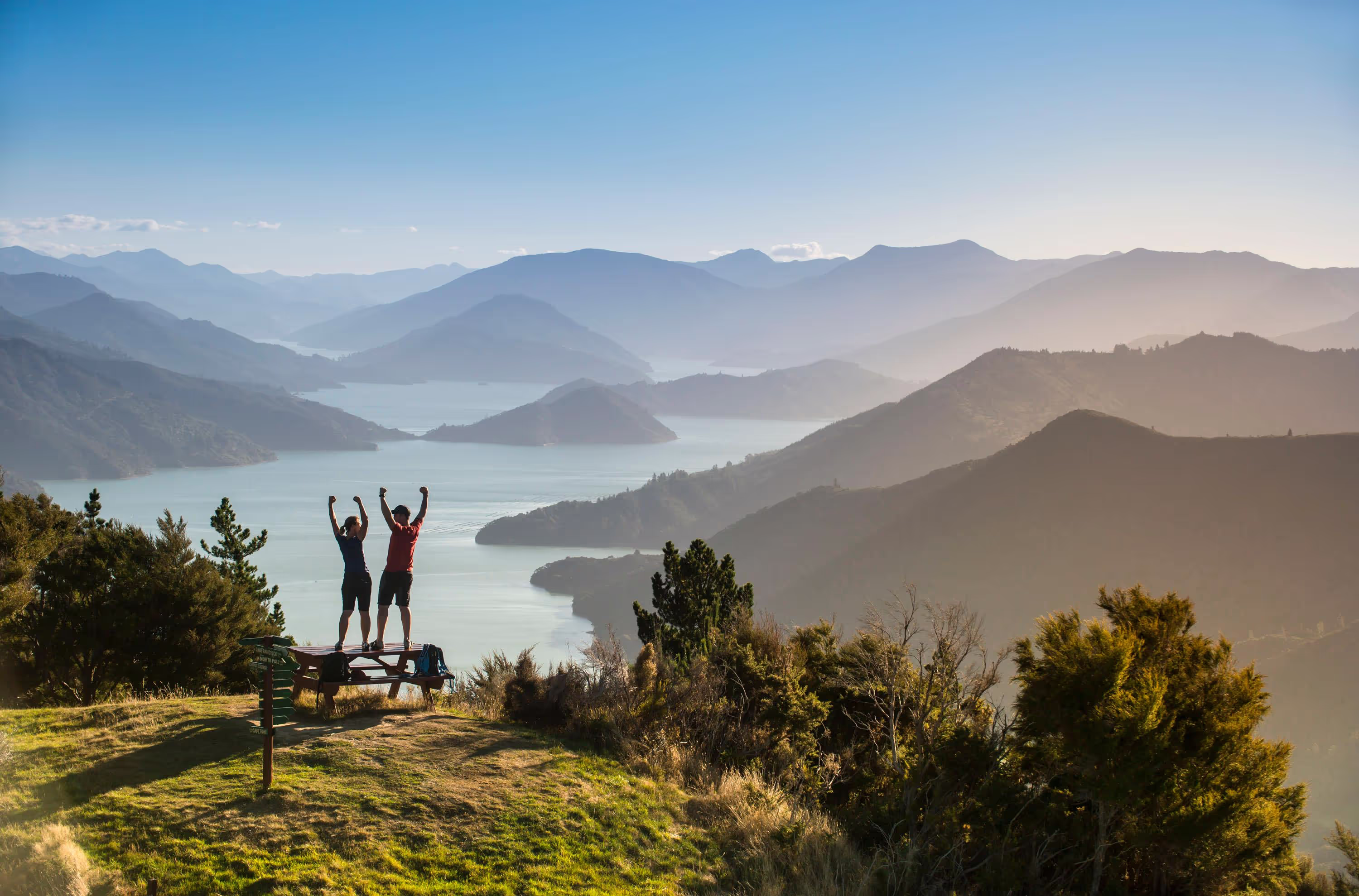 Two people raising arms on a mountaintop overlooking a lake and hills at sunset.