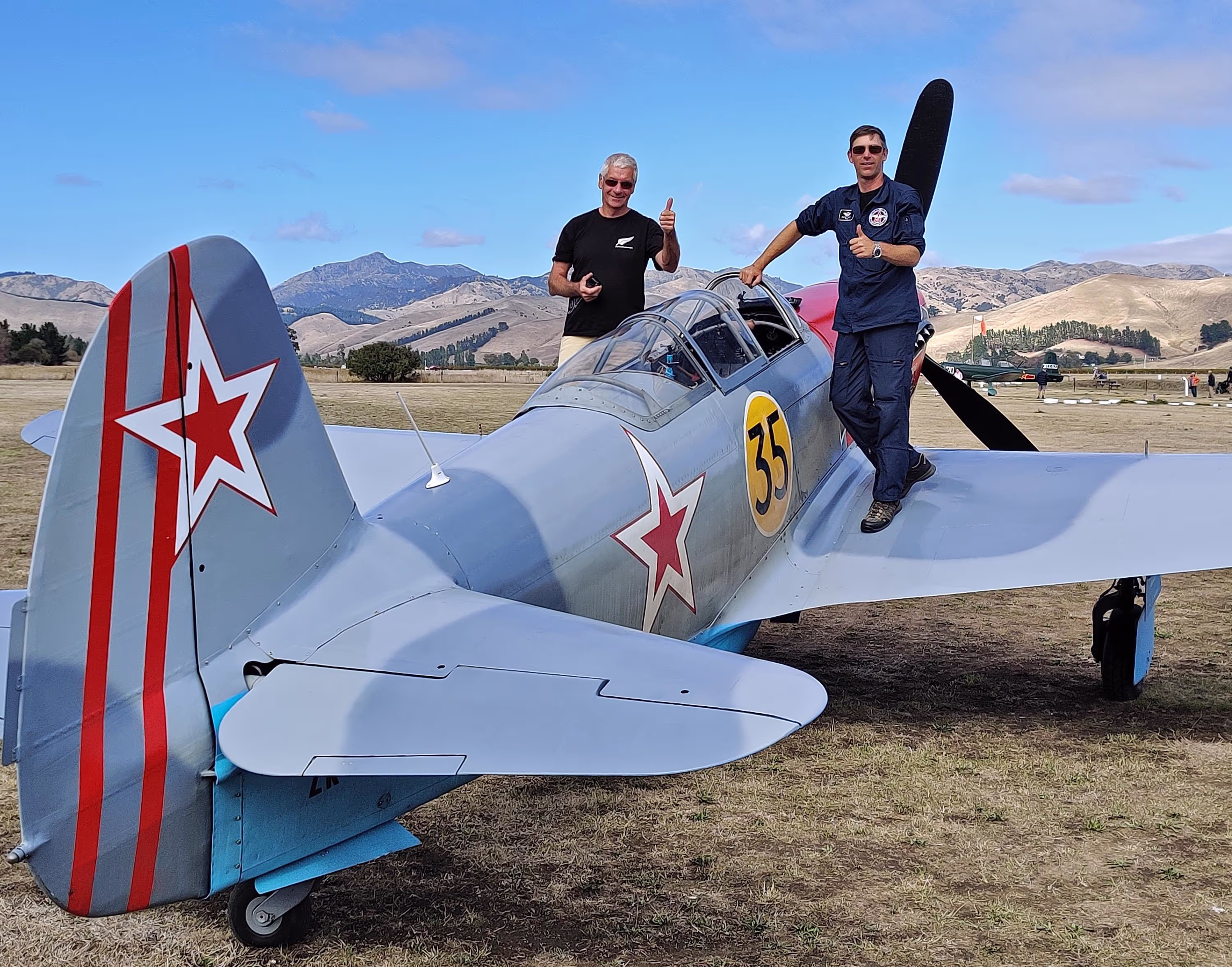 A vintage fighter plane with red star insignia on the tail parked outdoors with mountains in the background.