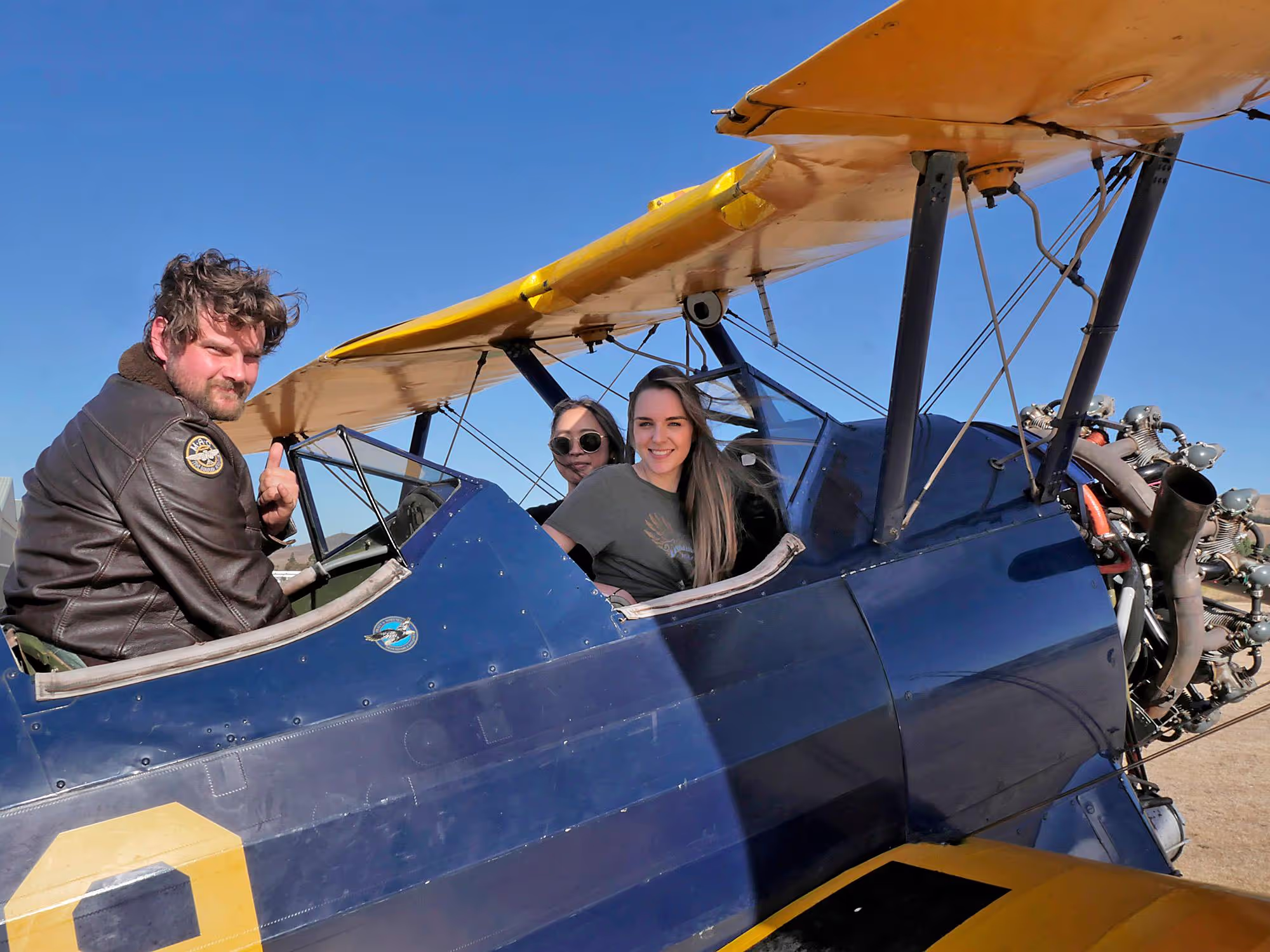 Two individuals in a vintage biplane, clear skies, one wearing a leather jacket.