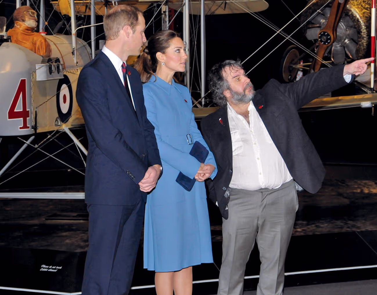 Three individuals in front of a vintage aircraft, one gesturing to the left.
