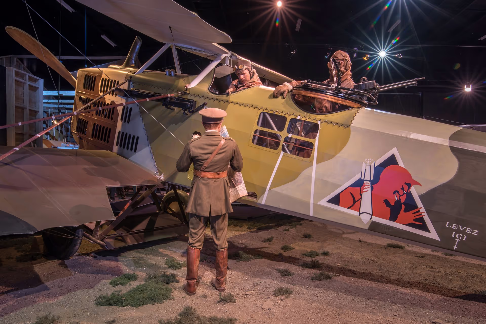 A person in vintage military attire looking at a historical biplane with figures inside at a museum exhibit.