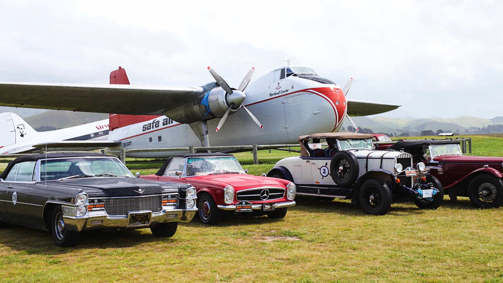 Red vintage fire engine and classic cars parked in a row at Omaka, with visitors and hangars behind.