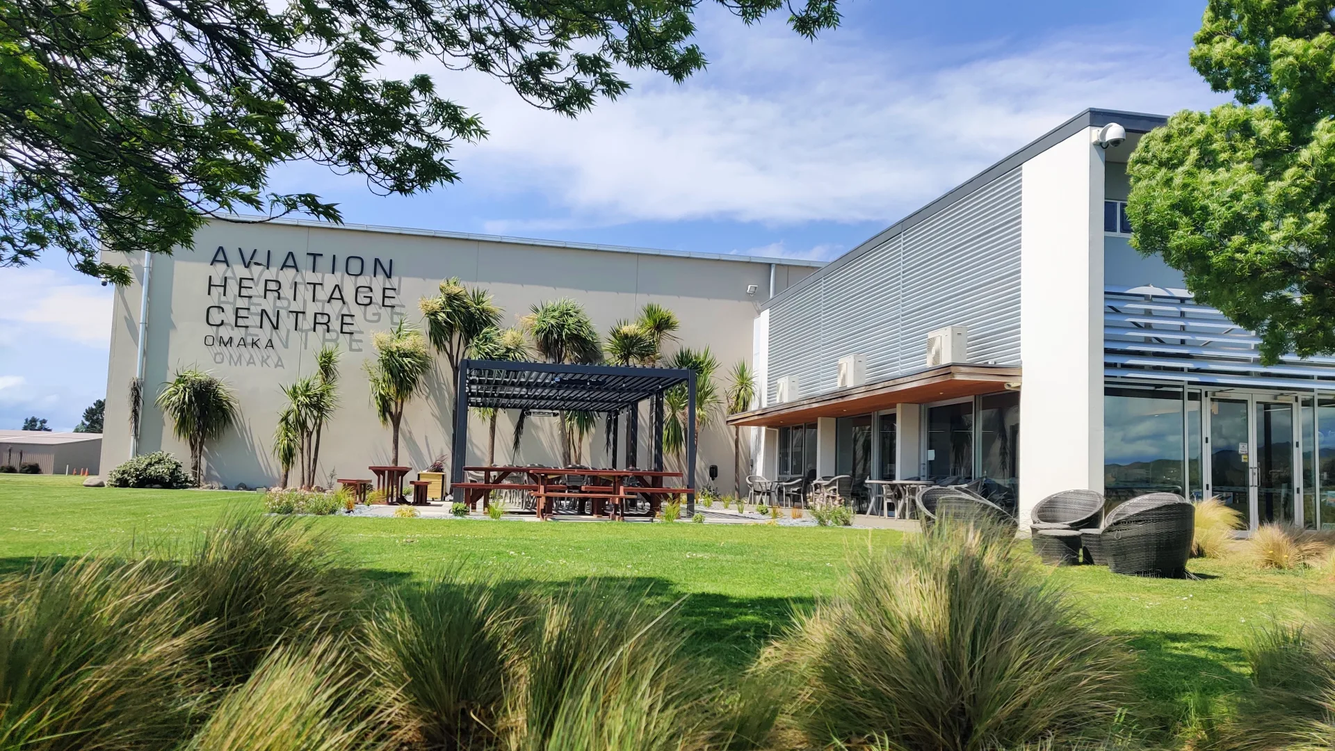 Exterior of the Aviation Heritage Centre in Blenheim, showing the museum building, lawn, outdoor seating area, native planting, and clear blue sky.