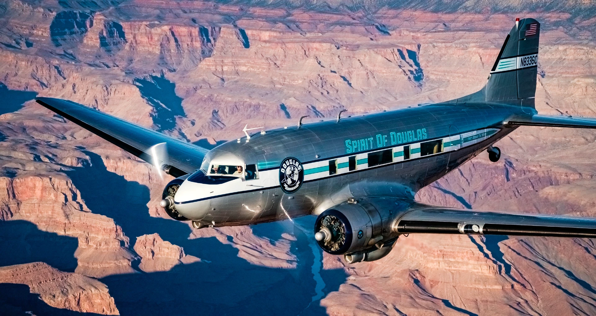Spirit of Douglas DC-3 flying above the Grand Canyon, with its polished silver fuselage and distinctive teal markings visible against the rugged landscape.
