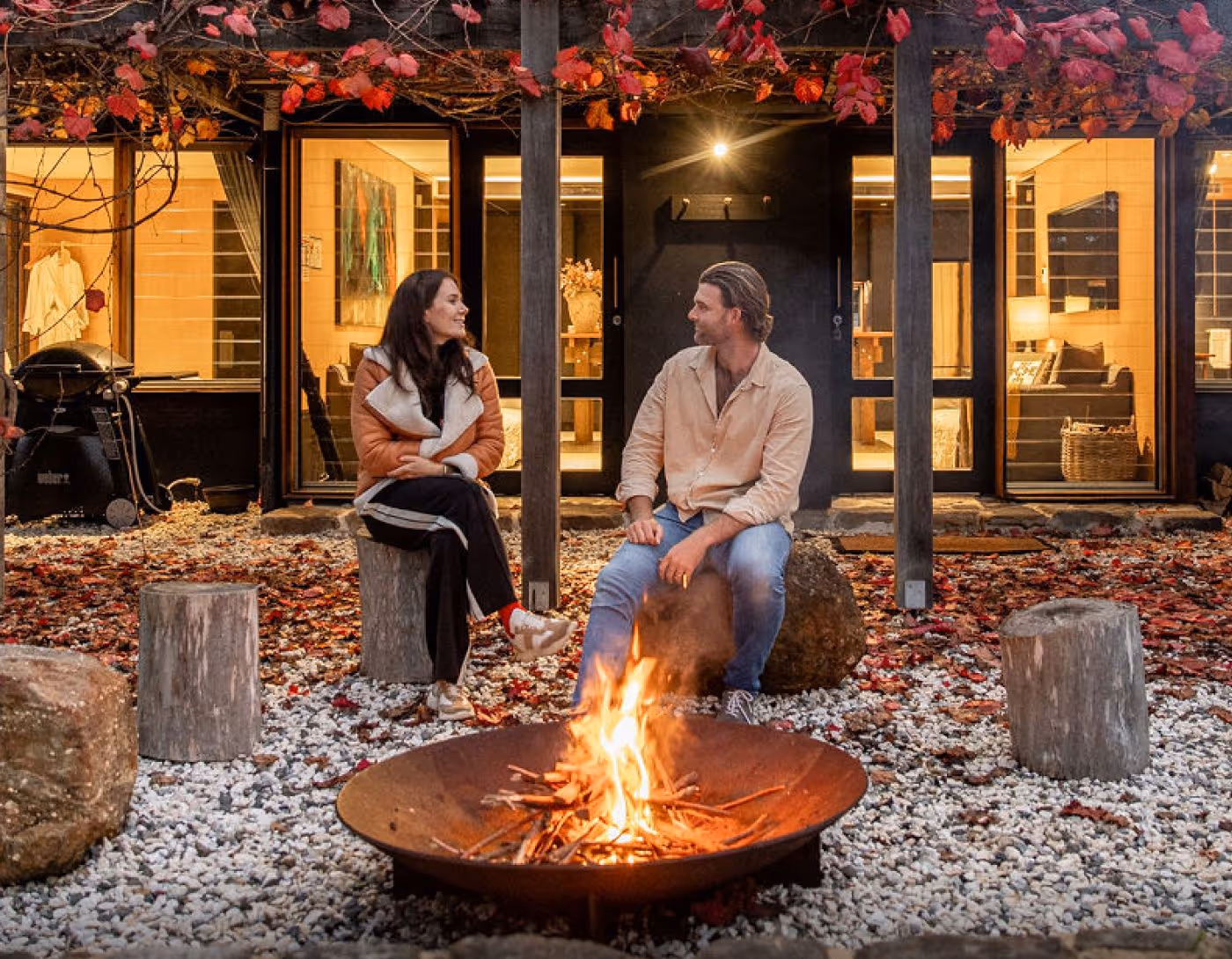 Couple sitting on Barranca's log stools around a fire pit outside a modern house with warm interior lighting and red autumn leaves overhead.