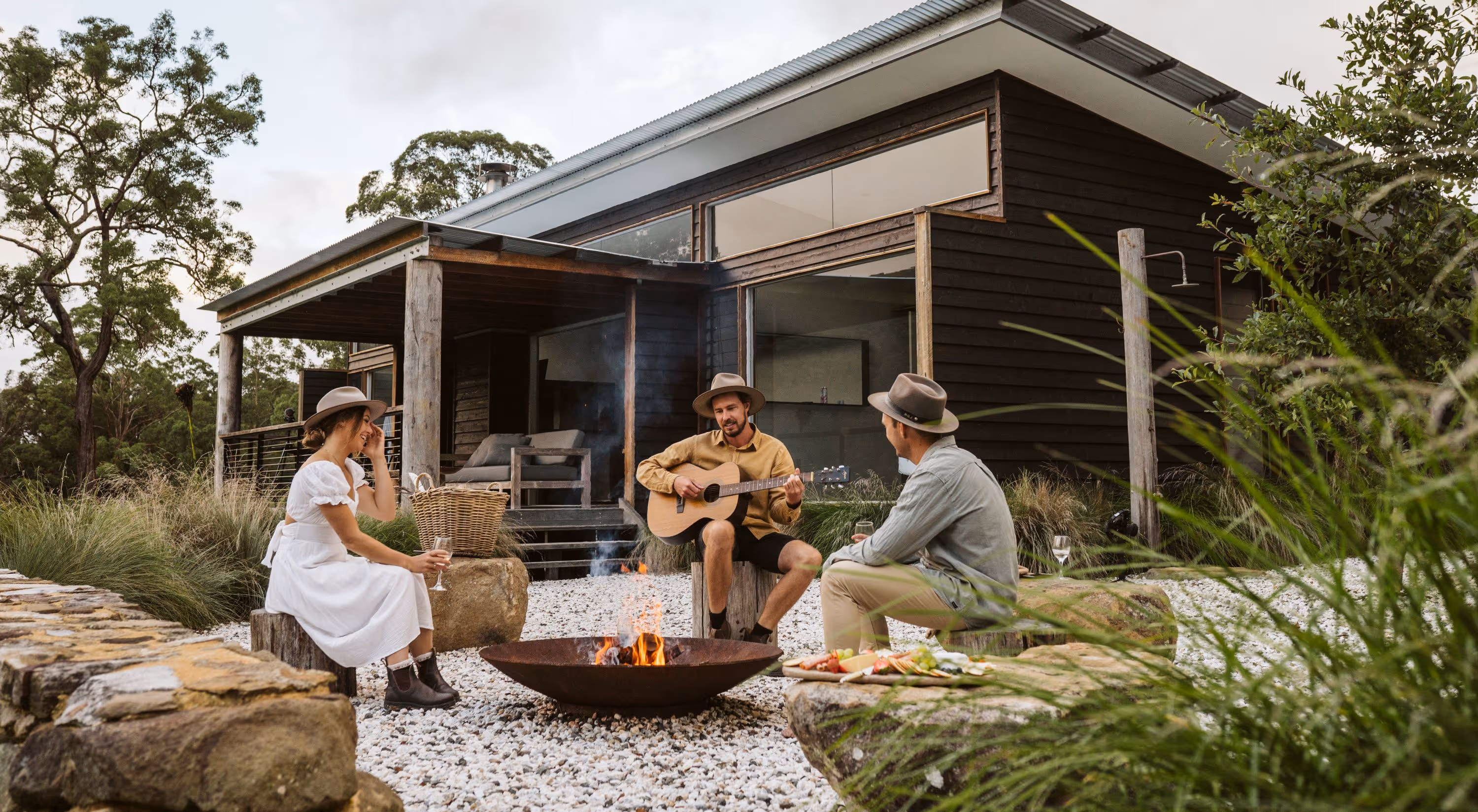 Three people wearing hats sitting around a fire pit outside a Barranca's modern wooden cabin, with one playing a guitar and others holding wine glasses.