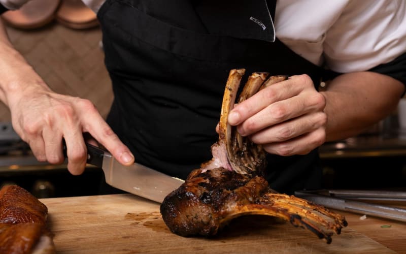 Chef carving roasted meat from a rack of ribs on a wooden cutting board