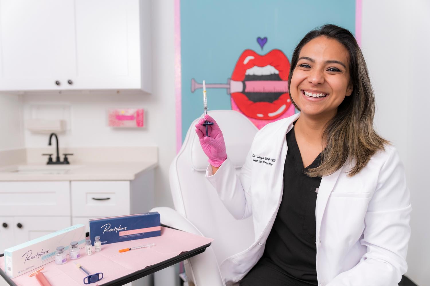 A healthcare professional in a white lab coat and pink gloves holds up a medical syringe in a bright, modern clinical setting. She smiles directly at camera with a colorful poster featuring red lips visible on the blue wall behind her, while Resty...