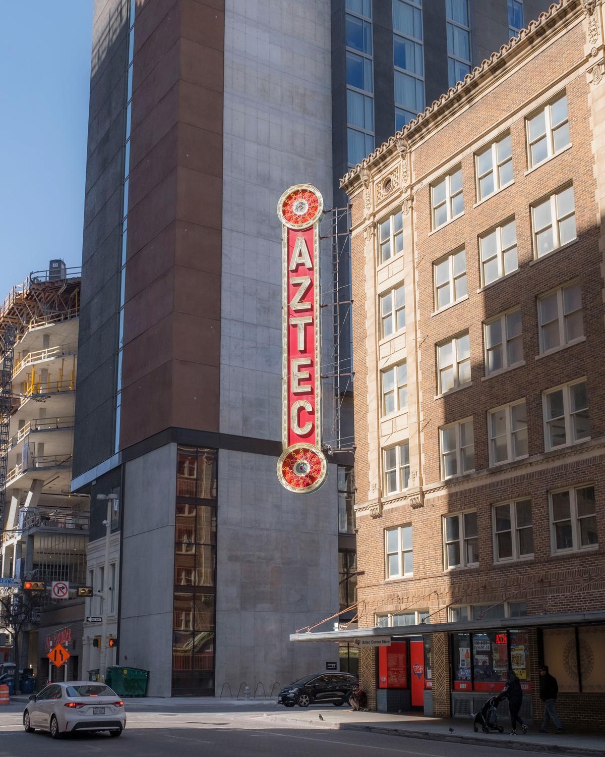 A tall vertical vintage Aztec theater sign mounted on a modern grey concrete building facade, positioned between contemporary architecture and a historic brick building with ornate detailing. The red and white illuminated sign features decorative ...