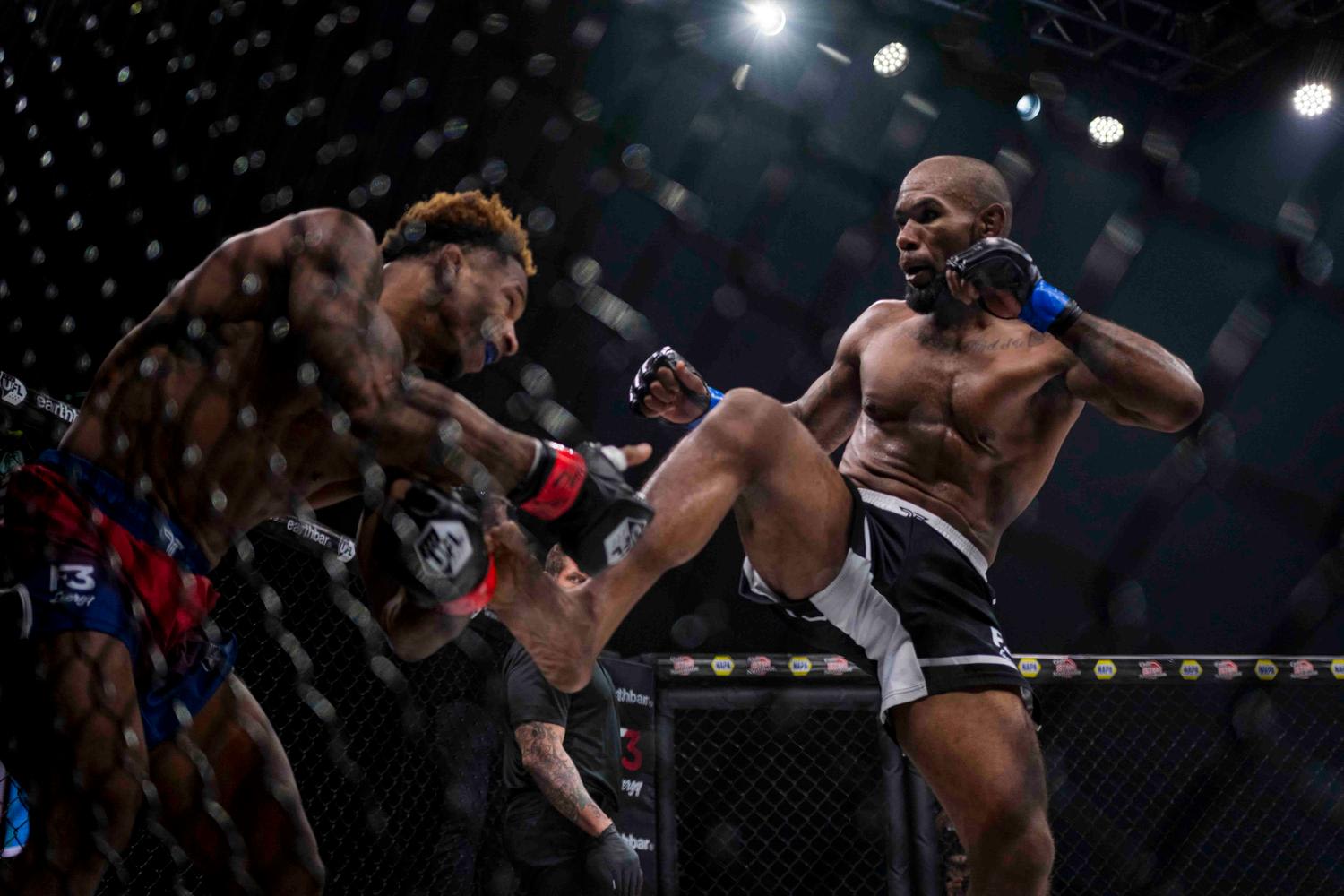 Two MMA fighters in an octagon cage during an active match, with one fighter executing a high kick while the other recoils. The scene captures intense athletic action under dramatic arena lighting with visible crowd and sponsor signage in the back...