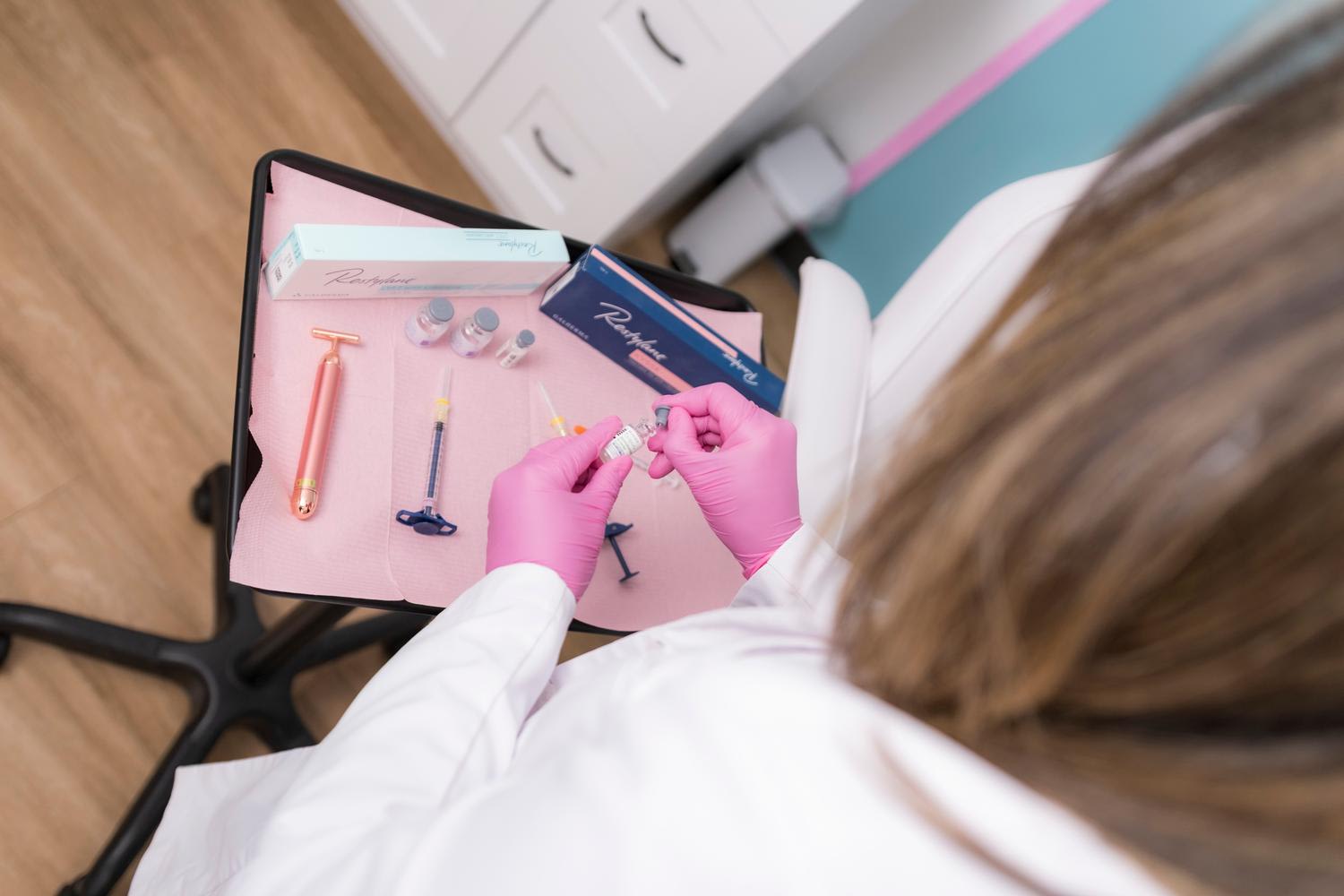 Overhead view of a beautician or aesthetician wearing pink gloves preparing cosmetic or medical beauty equipment on a pink tray, with various tools including syringes, applicators, and product packaging visible. The practitioner is seated at a cle...