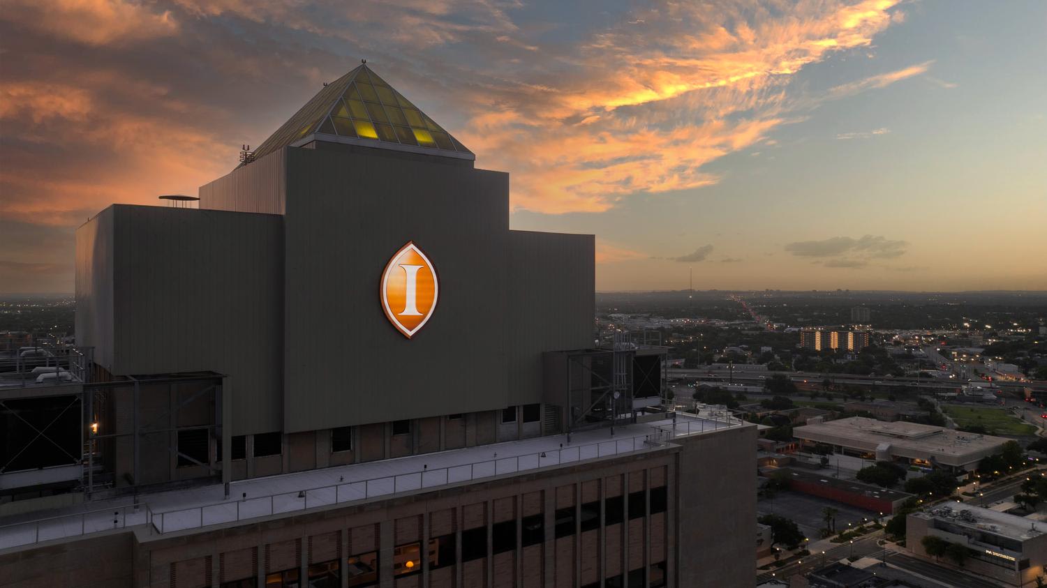 Wide aerial view of a modern multi-story institutional building with dark geometric architecture featuring a distinctive triangular rooftop with illuminated yellow accent windows and a prominent circular logo with the letter 'I'. The building sits...