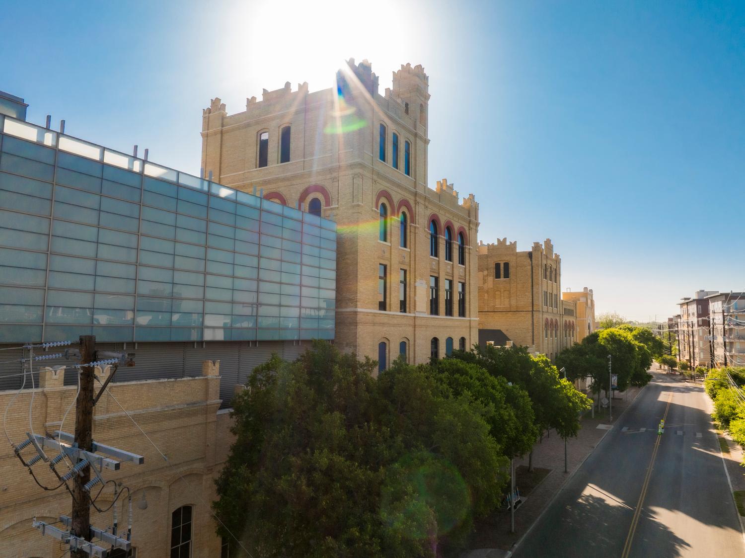 Wide-angle view of a historic brick building with ornamental crenellations and colorful arched windows, featuring modern glass addition on the left, with a sun flare creating lens effects across the facade. Tree-lined street and contemporary build...
