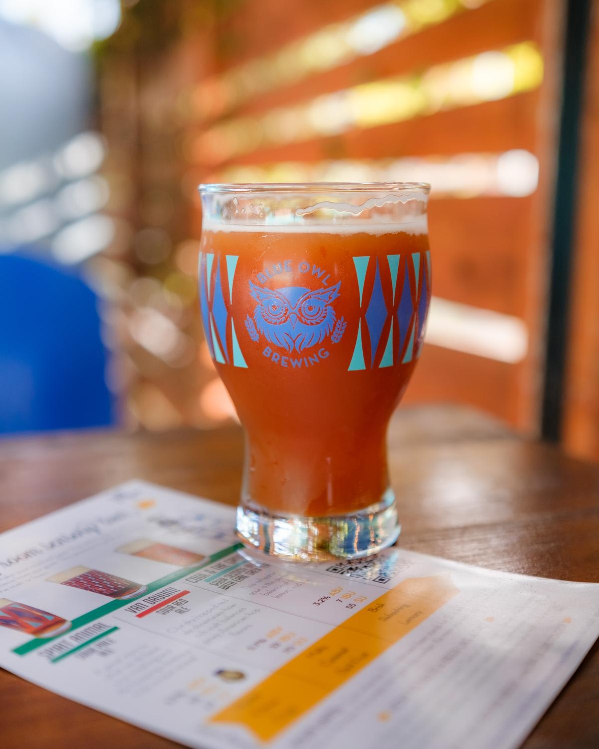 A tulip-shaped beer glass with amber-colored craft beer and foam head sits on a wooden table with printed menu materials beneath it. The glass features Night Owl Brewing branding with an owl logo and geometric pattern in blue, while warm orange to...