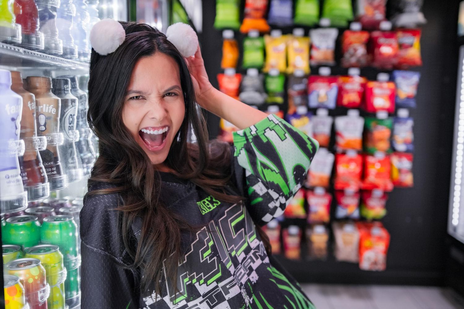 A young woman in a black gaming-themed shirt with green pixelated graphics holds a matching pixelated foam pickaxe while wearing white pom-pom headband ears, making an excited expression in a convenience store with beverage coolers and colorful sn...