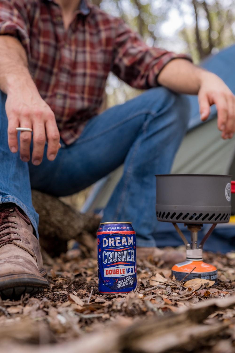 A can of Dream Crusher Double IPA beer is positioned on a leaf-covered ground next to a portable camping stove with a black pot. A person in plaid shirt, jeans, and hiking boots sits relaxed in the background, creating an outdoor recreational scene.