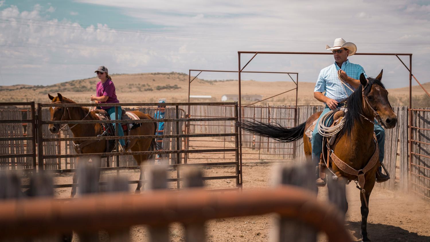 Two riders on horseback in western attire navigate a dusty rodeo arena with metal gate structures, one rider in a purple shirt on a brown horse in the background, another in a light blue plaid shirt and cowboy hat on a bay horse in the foreground ...