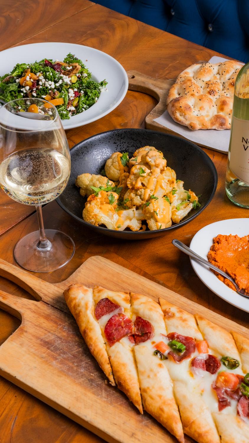 Overhead flat-lay composition of a restaurant table featuring white wine, multiple small plates including kale salad, buffalo cauliflower in a dark bowl, flatbread pizza slices on a wooden board, hummus, and seasoned naan bread arranged on a warm ...