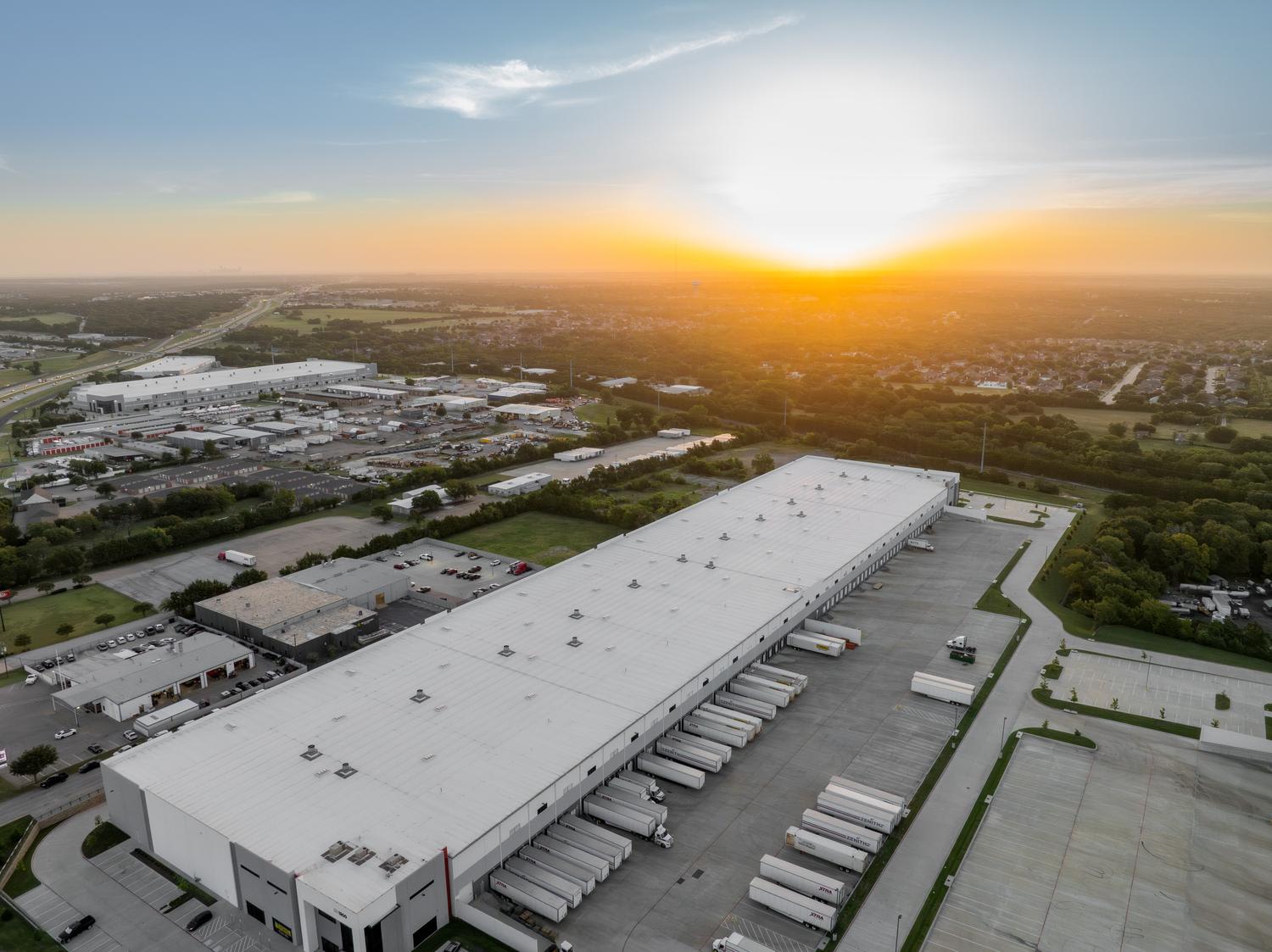 Aerial view of a large industrial warehouse and distribution center with multiple loading docks and semi-trucks parked alongside the building. The facility sits within a suburban industrial park at sunset, with additional commercial buildings and ...